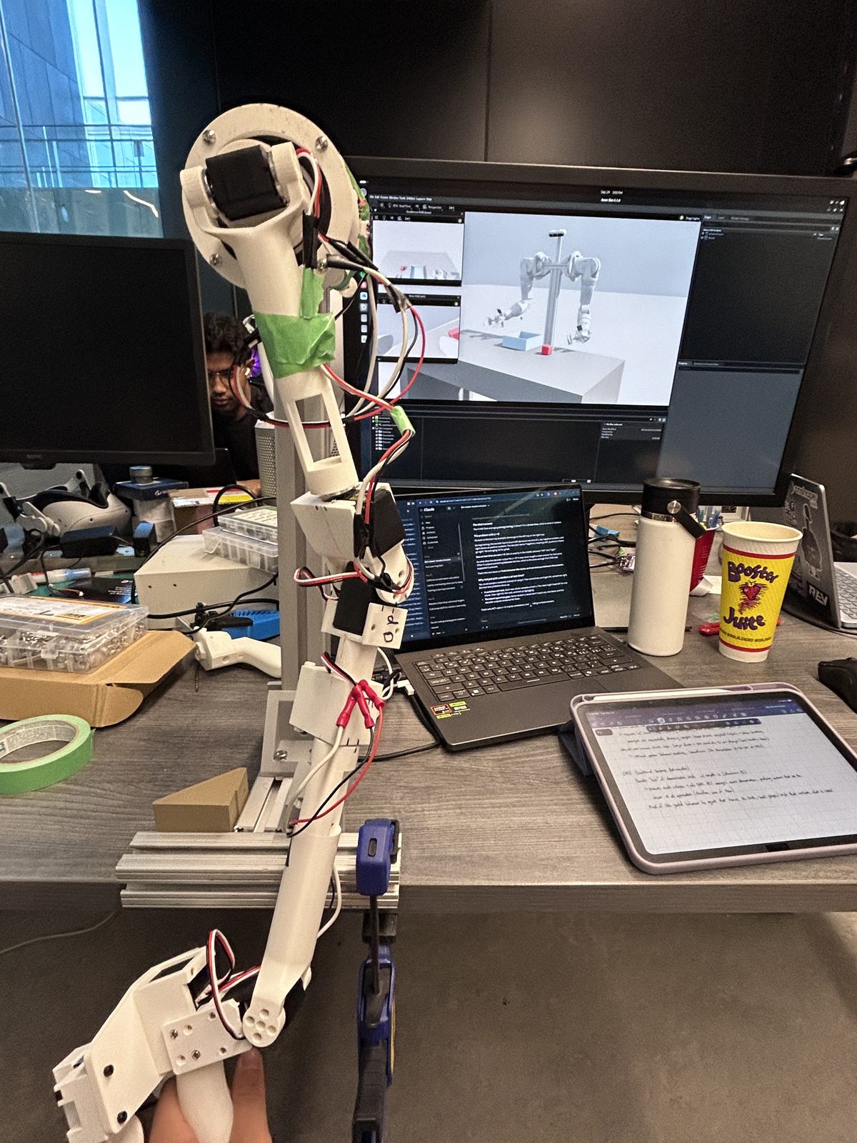 A 3D-printed leader arm clamped to a desk, with the Pioneer arm simulation on the monitor behind it